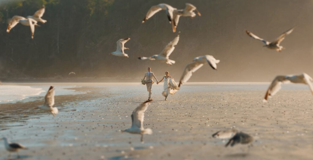 A couple runs holding hands on the beach as birds fly around them.