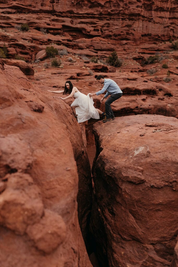 The groom helps the bride cross two rocks in her wedding dress.