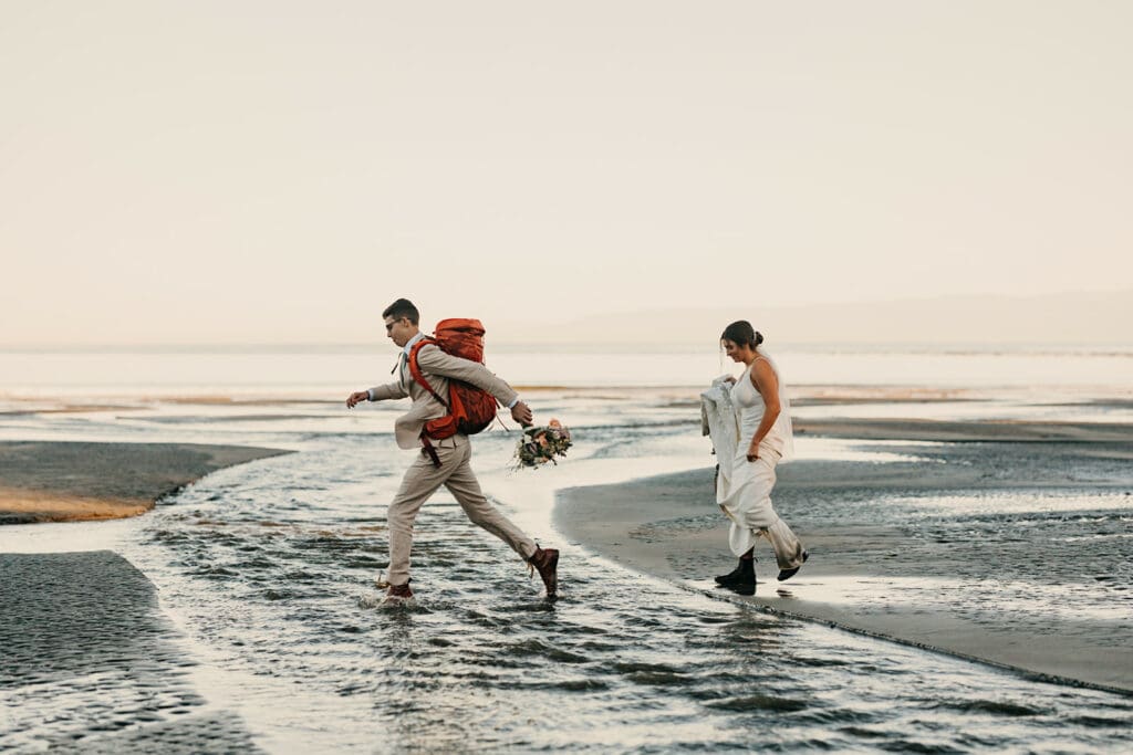 The groom and bride cross a river flowing on the beach shore.