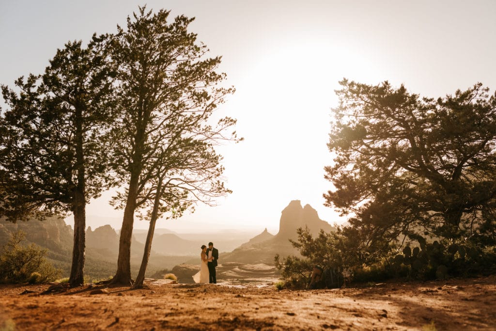A couple shares their first dance overlooking Sedona by large trees with the sun setting behind them.