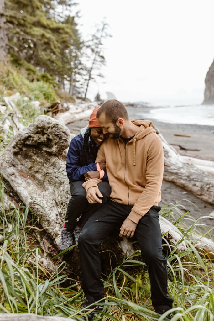 A couple cuddles and smiles while on a log on the beach.