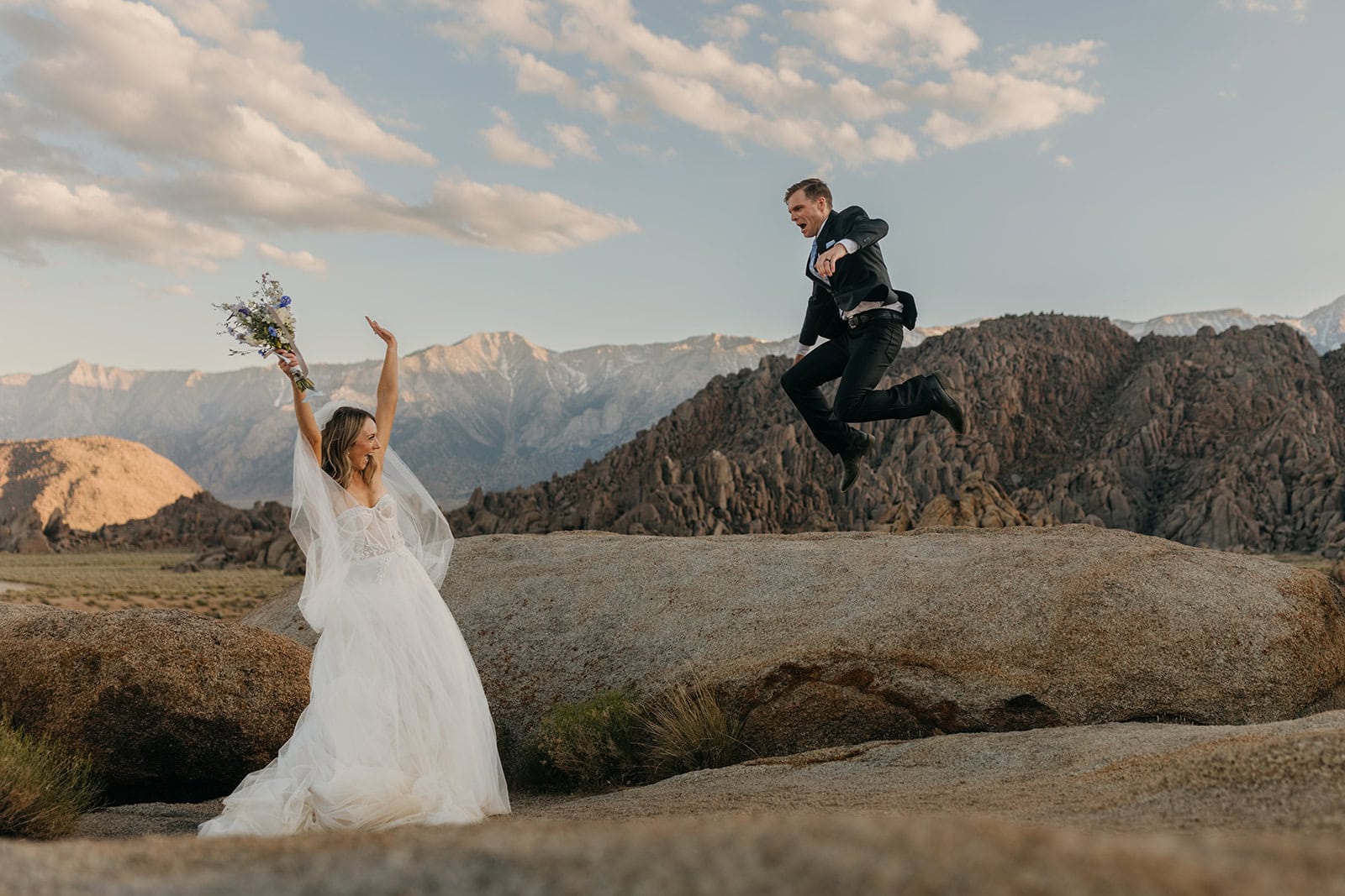 A groom jumps off of a rock while the bride celebrates.