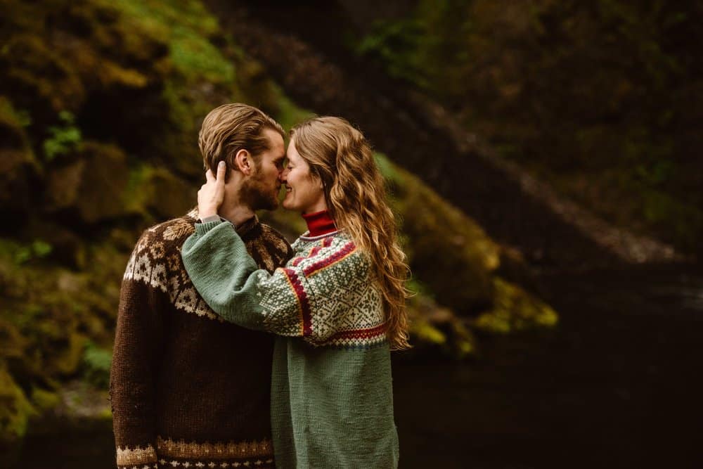 A couple in Nordic sweaters standing in front of a mossy wall together.