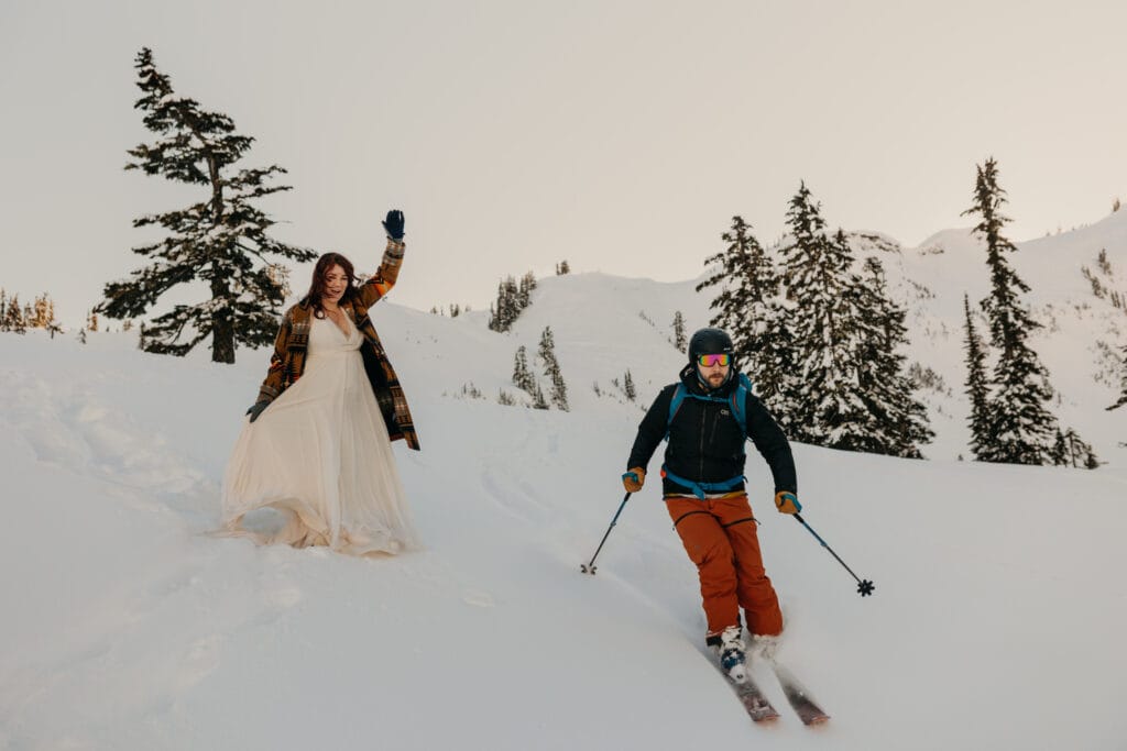 The groom skis past the bride on this snowy mountain during a winter wedding.