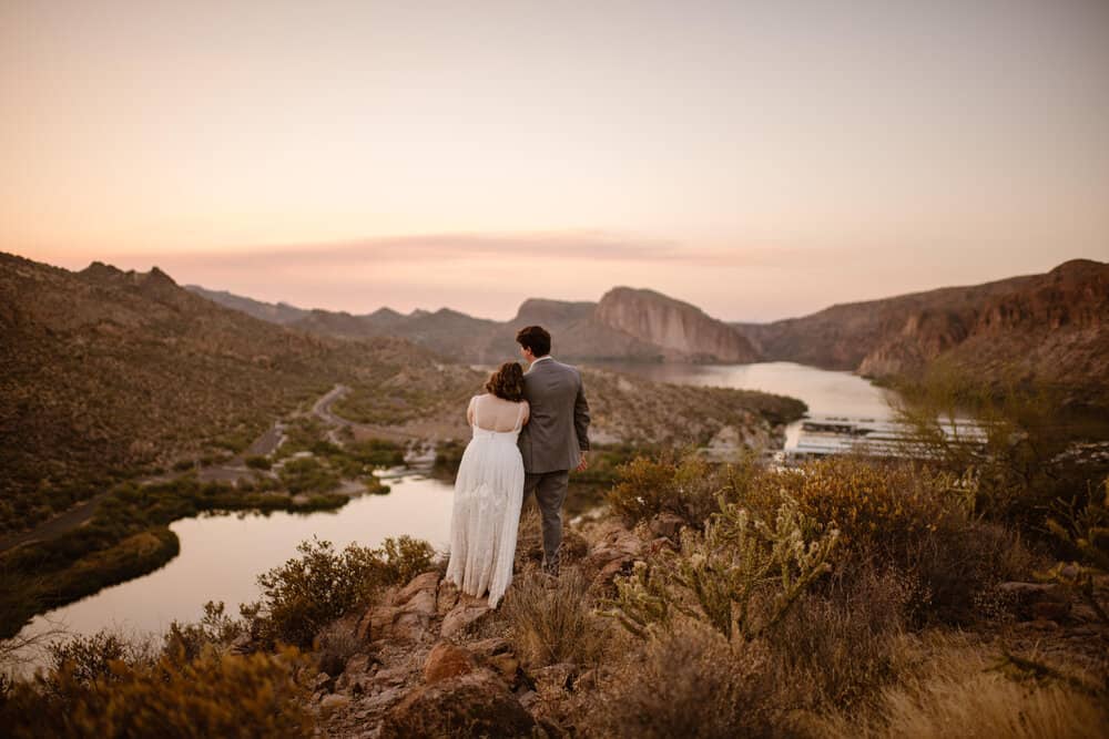 A bride leans on her grooms shoulder as they watch the sunset together.