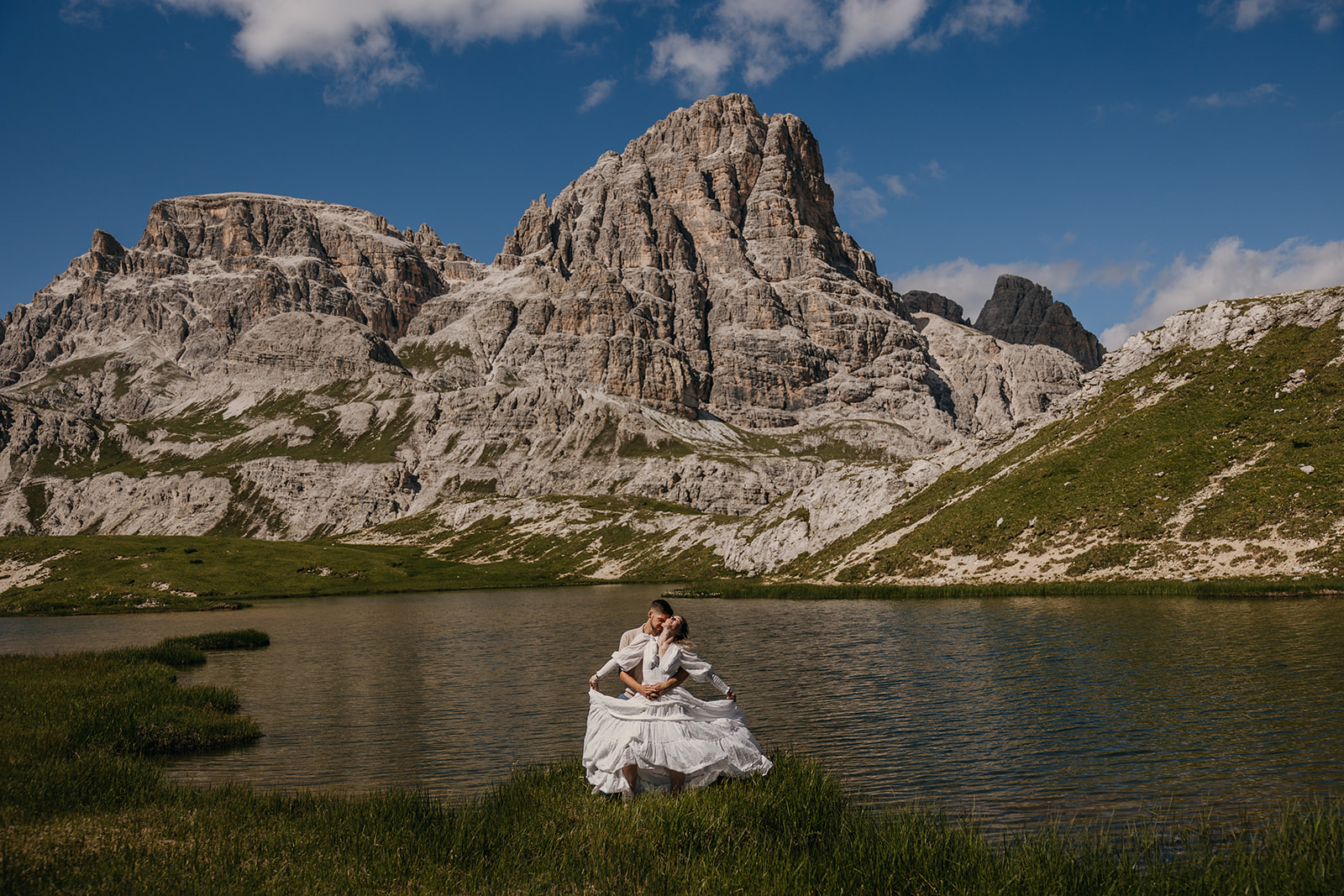 A couple stands in the sunshine together by a lake in the Dolomites. 