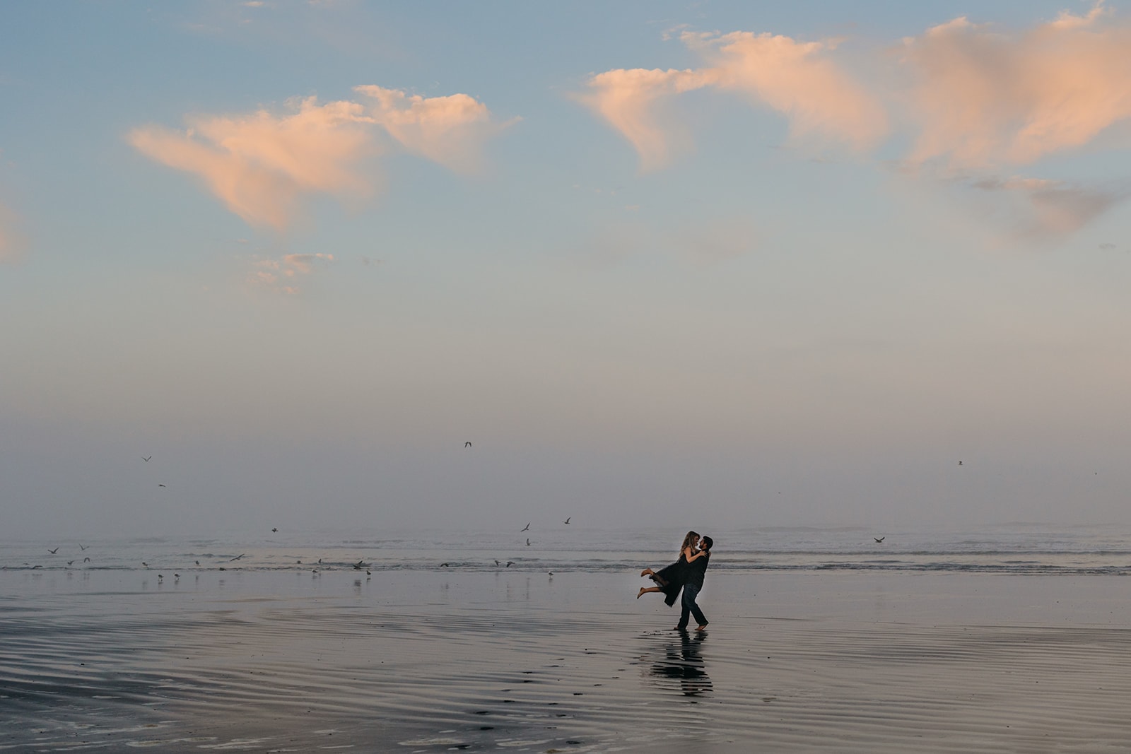 The groom swings the bride around on the beach in the morning light. 