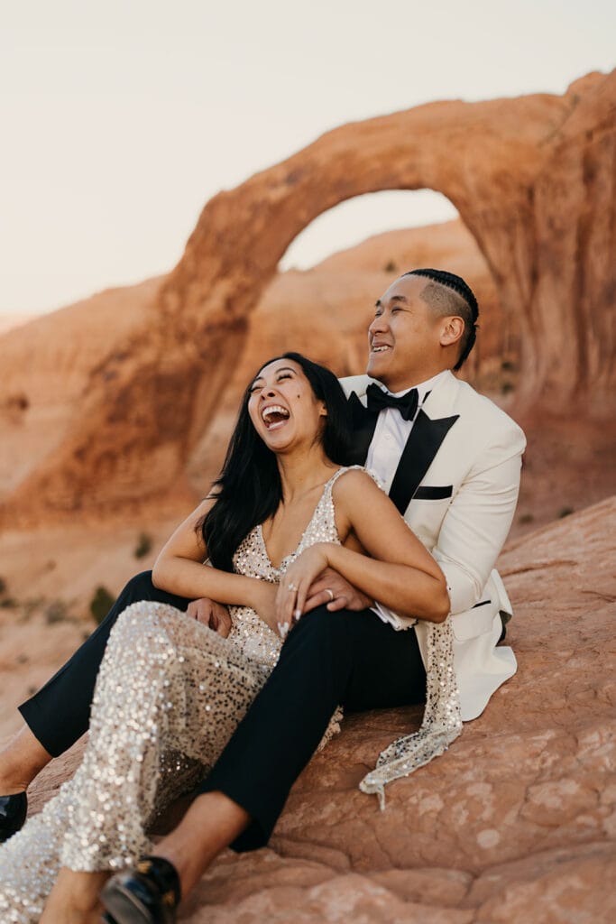 The couple sits together in the desert laughing in beautiful attire.