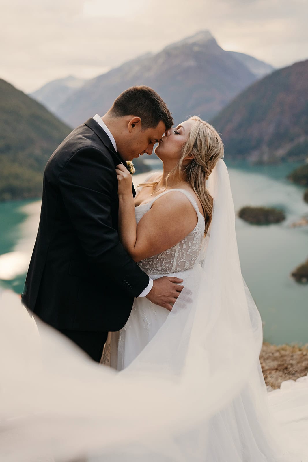 A bride kisses her grooms forehead in the mountains overlooking a lake.