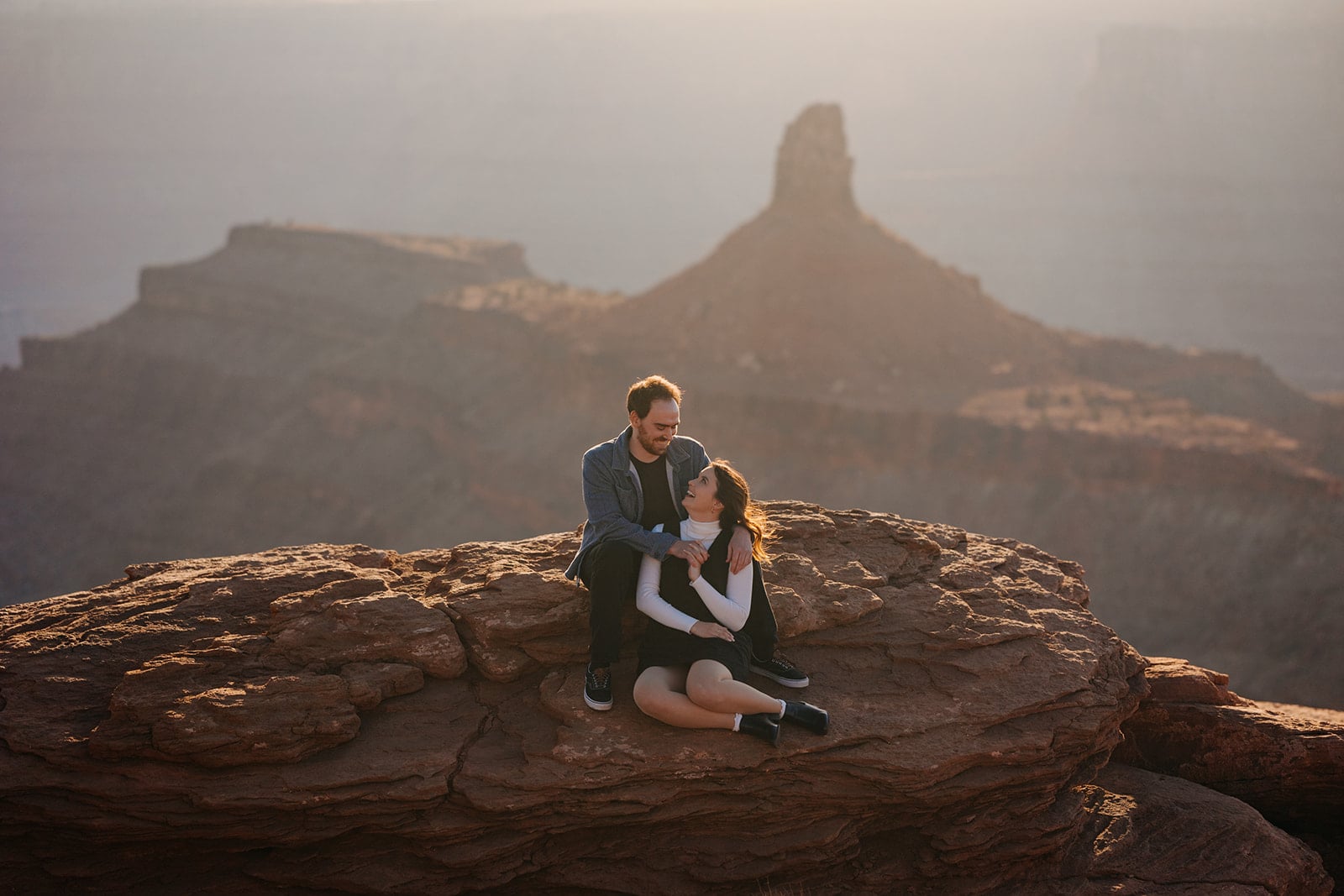 A man and woman sit together on a high up ledge at sunset in Moab