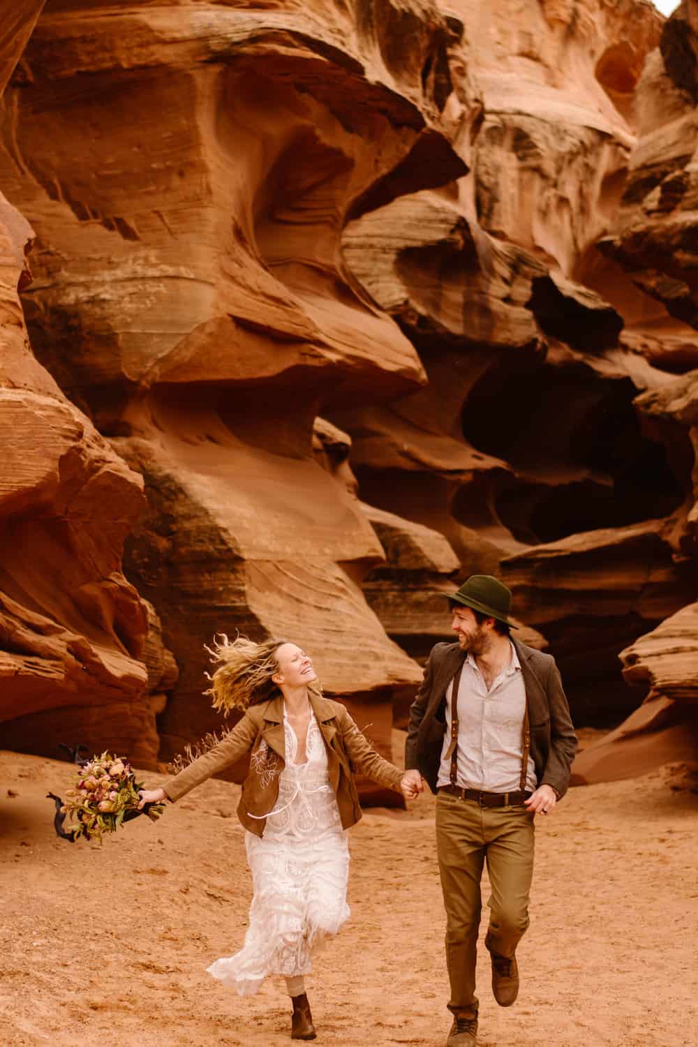 A bride and groom run through a slot canyon together holding hands and smiling.