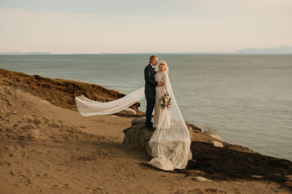A bride looks out at the ocean while a groom admires her and the wind catches her veil.
