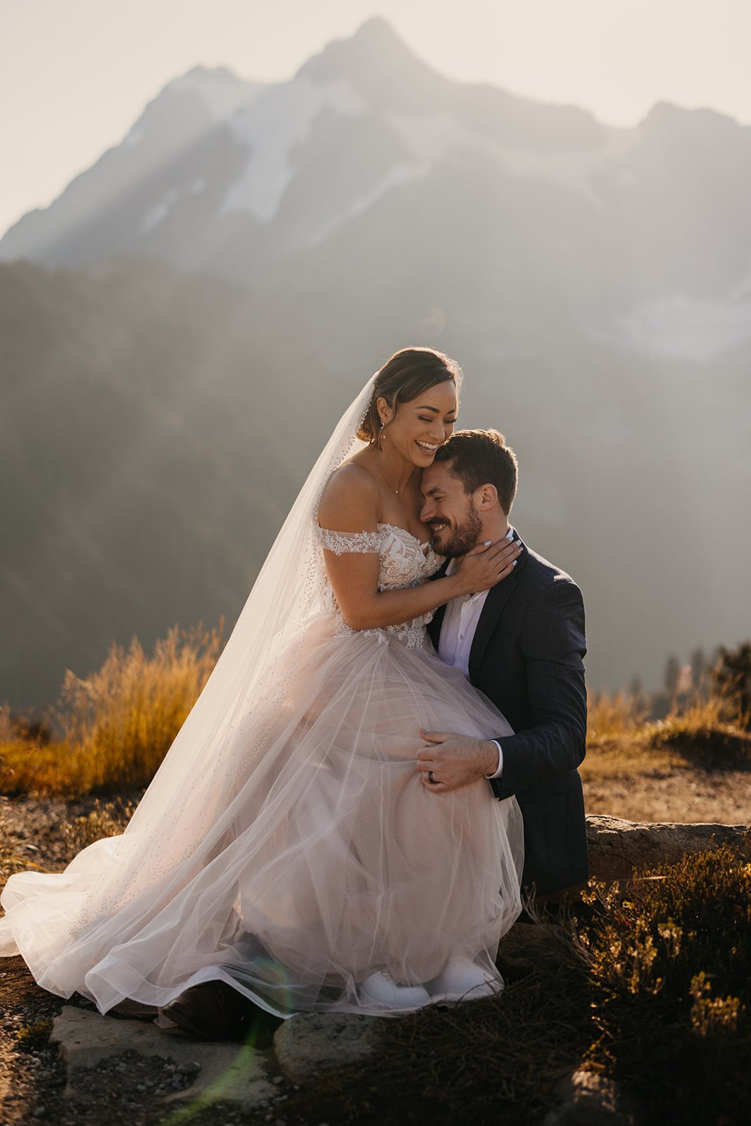 A bride embraces her groom smiling as they sit in the mountains together.