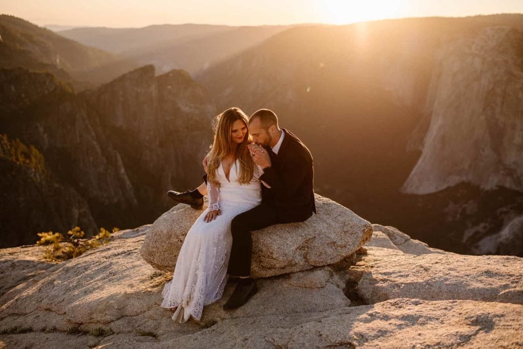 A groom kisses a brides hands as the sun sets behind them.